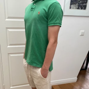 Ralph Lauren piké - Säljer en klassisk grön pikétröja från Polo Ralph Lauren i storlek M. Tröjan har korta ärmar, krage och knappar framtill samt den ikoniska röda loggan broderad på bröstet. Perfekt för en stilren och avslappnad look. Modellen är 183 cm och nypriset är cirka 1200 kr.
