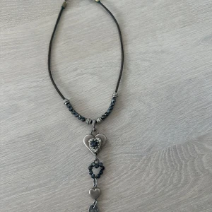 Halsband med hjärtan och svarta pärlor - Unikt halsband med svart snöre, dekorerat med silverfärgade detaljer och svarta pärlor. Hänget består av flera hjärtan och gnistrande stenar. Perfekt accessoar för att lyfta din outfit.
