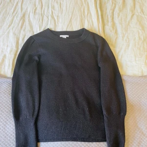 Svart stickad tröja med rund hals, lite glittrig - En svart stickad tröja med rund halsringning och långa ärmar. Tröjan har en klassisk, enkel design och sitter normalt på kroppen. Perfekt för lager-på-lager och passar till många olika outfits.