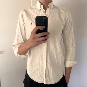 Vit skjorta från Ralph Lauren - Klassisk vit skjorta från Ralph Lauren med broderad logga på bröstet. Skjortan har lång ärm, knappar framtill och en tidlös passform. Perfekt för dig som gillar stilrena plagg med ikonisk touch. Lite märke under ärmarna men inte märkbart vid användning!