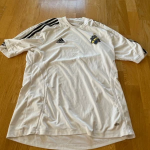 Vit AIK fotbollströja från Adidas - Säljer en vit AIK fotbollströja från Adidas med svarta ränder på axlarna och AIK-logga på bröstet. Tröjan har korta ärmar och detaljer i guld, inklusive 'A.I.K.' i nacken. Perfekt för match eller träning. Officiell matchtröja från 2000-tal. 