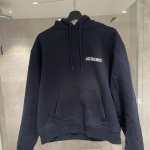 Marinblå hoodie från Jacquemus - Säljer en svart hoodie från Jacquemus med vit logga på bröstet. Klassisk modell med huva och känguruficka framtill. Perfekt för en avslappnad och stilren look.