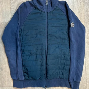 Mörkblå quiltad jacka från Colmar - Mörkblå jacka från Colmar med quiltad framsida och baksida samt mjuka ärmar. Jackan har dragkedja framtill och en liten logotyp på bröstet. Perfekt för kyliga dagar och enkel att matcha med det mesta.