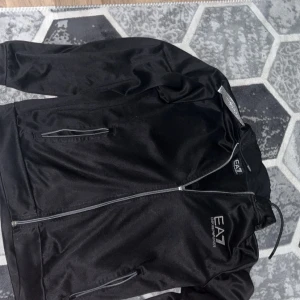 Svart jacka med dragkedja från EA7 Emporio Armani - Säljer en svart jacka från EA7 Emporio Armani med huva och dragkedja framtill. Jackan har två sidofickor med dragkedja och EA7-logga på bröstet. Perfekt för en sportig och stilren look.