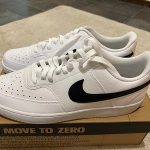 Nike vita sneakers med svart swoosh - Fräscha vita Nike sneakers med klassisk svart swoosh på sidan. Skorna har snörning, rund tå och platt sula. Perfekta för en clean och sportig look. Modellen är enkel men stilren och passar till det mesta.