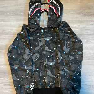 BAPE hoodie - Säljer en svart camo hoodie från BAPE med hajmotiv på huvan och broderade bokstäver på sidan. Hoodien har dragkedja hela vägen upp och coola detaljer i blått, vitt och grått. Perfekt för dig som gillar streetwear och vill sticka ut.