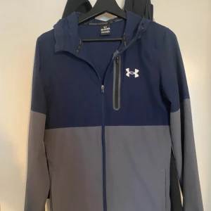 Under Armour jacka  - Säljer en snygg tvåfärgad vindjacka från Under Armour i blått och grått. Jackan har huva, dragkedja framtill och en bröstficka med dragkedja. Perfekt för träning eller vardag. Storleken är XS men sitter som en S. 