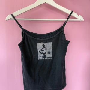 Svart linnetopp med tryck 'Little Sister' - Svart linnetopp med tunna axelband och tryck på bröstet där det står 'Little Sister' tillsammans med en bild. Perfekt för en avslappnad och cool stil.