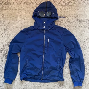C.P. Company jacka från 2000-talet - Säljer en blå vindjacka från C.P. Company med avtagbara goggles och luva. Storlek 54, passar som L/XL. Vet inte exakt namn på modellen men har hittat jackan på andra sidor som säger att den är från 2000-talet. Bra skick! Hör av er för fler bilder!