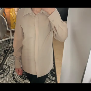 Beige skjorta med klassisk krage - Stilren beige skjorta med klassisk krage och dolda knappar framtill. Skjortan har långa ärmar med knapp vid manschetten och rak passform. Perfekt för en clean och enkel look.