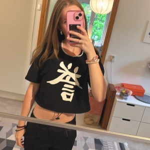 Svart croppad t-shirt med vitt tryck - Svart croppad t-shirt från new yorker med stort vitt tryck av ett tecken på framsidan. T-shirten har korta ärmar och rund halsringning. Det sitter en snodd i midjan så man kan dra åt om man vill.🩷