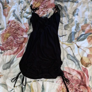 Svart kortklänning med knyt från Missguided - Säljer en svart kortklänning från Missguided med smala axelband som knyts och draperad urringning. Klänningen har knytband i sidorna nertill för justerbar längd och passform. Perfekt för dig som gillar en stilren och trendig look😍