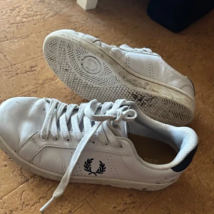 Vita sneakers från Fred Perry - Säljer ett par klassiska vita sneakers från Fred Perry med svart logga på sidan och svart detalj vid hälen. Skorna har snörning och rund tå, samt platt sula. Perfekta för en stilren och sportig look.skorna är renare än vad det ser ur