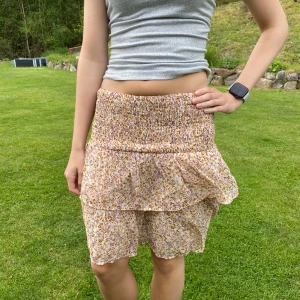 Blommig volangkjol från ADPT - Helt ny och oanvänd! Supersöt kort blommig kjol från ADPT med volanger och smock i midjan. Kjolen har ett ljust beige tyg med små lila och gula blommor, perfekt för en somrig look. Luftig och lätt, passar perfekt till varma dagar. Går att vika ner om man vill ha en kortare modell. 