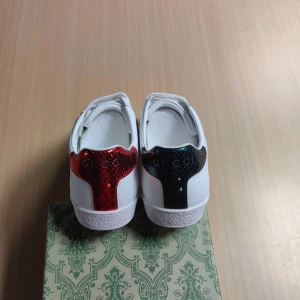 Gucci vita sneakers med orm och färgdetaljer - Säljer ett par vita sneakers från Gucci med klassiska gröna och röda ränder på sidorna samt broderad orm. Baktill har ena skon röd och andra skon grön metallic-detalj. Skorna har snörning och platt sula.