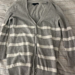 Grå och vit randig kofta från Tommy Hilfiger  - Säljer en grå kofta från Tommy Hilfiger med vita horisontella ränder. Koftan har knappar framtill, v-ringning och långa ärmar. Perfekt att slänga över en t-shirt eller linne för en avslappnad look.