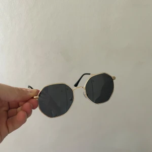 Svarta solglasögon med guldiga bågar - Snygga solglasögon med svarta, åttakantiga glas och tunna guldiga metallbågar. Skalmarna är svarta och har en stilren design. Perfekta för att ge din outfit en cool touch.