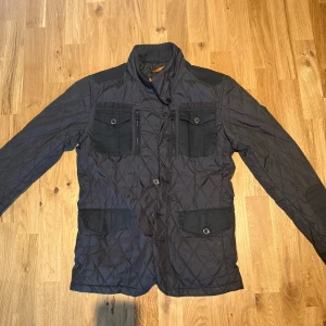 Svart quiltad jacka med fickor - Säljer en svart quiltad jacka med hög krage och fyra framfickor med lock och knappdetaljer. Jackan har lång ärm och stängs med både dragkedja och knappar. Perfekt för kyligare dagar.