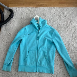 Turkos hoodie från Polo Ralph Lauren - Turkos hoodie med dragkedja och huva från Polo Ralph Lauren. Jackan har två fickor framtill och en broderad logga på bröstet. Perfekt för en avslappnad stil.