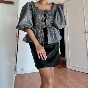 Glittrig silver topp med puffärmar från H&M - Säljer en glittrig silverfärgad topp från H&M med stora puffärmar och snörning framtill. Toppen har en peplum-liknande form och är perfekt för att sticka ut. Materialet är syntet och ger en lyxig känsla.