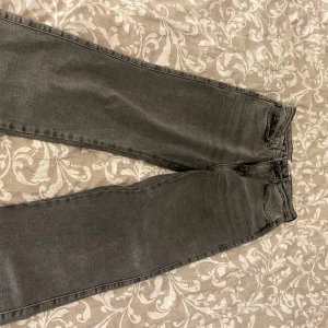 Gråa raka jeans från Madlady - Säljer ett par gråa jeans från Madlady med raka ben och hög midja. Klassisk femficksmodell med dragkedja och knapp framtill. Jeansen har en snygg tvättad look och passar perfekt till vardagsstilen. Ena remmen har gått av som bilden visar, därav säkerhetsnålen. Inget som syns när man har på dom.