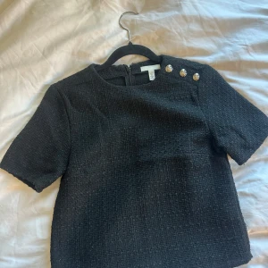 Svart kortärmad topp med dekorativa knappar - Stilren svart topp i strukturerat tyg med korta ärmar och tre dekorativa silvriga knappar på axeln. Toppen har rund halsringning och dragkedja i ryggen. Perfekt för en chic och klassisk look.