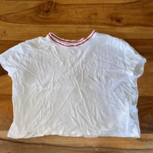 Vit croppad t-shirt med röd randig krage från H&M - Vit croppad t-shirt från H&M med korta ärmar och rund halsringning med röda ränder. Tillverkad i en mjuk bomulls- och modalblandning. Perfekt för en enkel och stilren look. En aning genomskinlig.