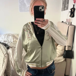 Grön satin jacka! - Super snygg ljusgrön bomberjacka i satin från Lindex med broderade blommor och fågel både fram och bak. Jackan har vita ärmar, fickor, dragkedja och ribbade muddar med gulddetaljer. Perfekt till allt! Önskade jag kunde andvända den men har så mycket jackor så den bara blir liggandes i gadroben!
