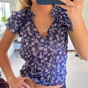 Blommig blus med volangärmar - Söt blus i mörkblått med ljusrosa och vita blommor. Blusen har korta volangärmar och v-ringning. 