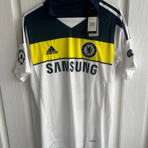 Chelsea bortatröja Torres 9 - Chelsea FC bortatröja med Torres 9 på ryggen. Tröjan är vit med mörkblå och gul detalj över bröstet, Adidas-logga och klubbmärke framtill samt Samsung som sponsor. Kortärmad modell med klassisk krage och Premier League-märke på ärmen.
