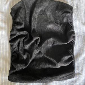 Svart glittrig tubtopp - Säljer en snygg svart tubtopp med glittrigt material. Toppen är axelbandslös men har tunna band som kan korsas i ryggen för extra stil. Perfekt för festliga tillfällen!