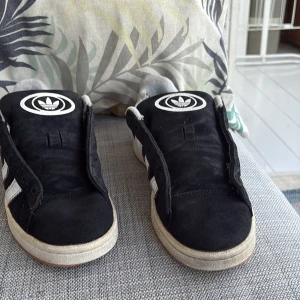 Svarta Adidas Campus sneakers - Säljer ett par klassiska svarta Adidas Campus sneakers med vita ränder och gummisula. Skorna har Adidas logga på plösen och texten 'CAMPUS' på sidan. Perfekta för dig som gillar retrostil och streetwear.