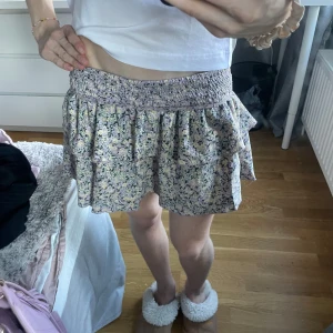 Blommig kortkjol med volang - Supersöt kortkjol med blommigt mönster i lila, beige och grönt. Kjolen har bred smock i midjan och volang nertill som ger en flowy känsla. Perfekt för varma dagar och enkel att styla med en enkel topp.