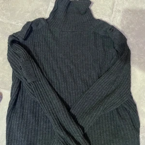 Mörkgrå ribbad polotröja med knappar - Säljer en mörkgrå ribbad polotröja med långa ärmar och snygga knappdetaljer på axlarna. Perfekt för kyligare dagar och enkel att styla till olika outfits. Stickad 