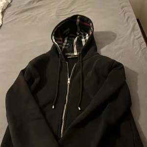 Svart hoodieZip från Burberry - Säljer en svart hoodiejacka från Burberry med klassiskt rutigt foder i svart, vitt och rött. Jackan har huva med dragsko och är helsvart på utsidan. Perfekt för dig som gillar stilrena plagg med lyxig känsla. Knappt använd då den är för liten för mig.