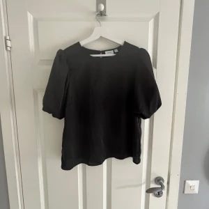 Svart blus med puffärmar från VILA - Säljer en svart blus från VILA med puffiga korta ärmar och rund halsringning. Blusen har en stilren design och knäppning bak i nacken. Perfekt för dig som gillar en enkel men trendig look.