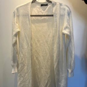 Vit stickad kofta - Säljer en lång, öppen vit stickad kofta med lång ärm. Koftan har en enkel och klassisk design utan knappar eller dragkedja, perfekt att slänga över en topp eller t-shirt. Passar till många olika stilar och tillfällen.