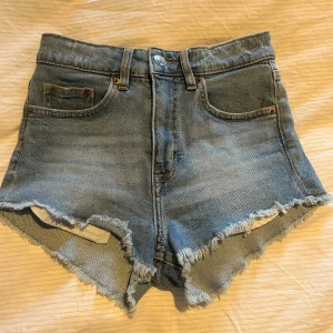 Blå jeansshorts med fransar - Säljer ett par klassiska blå jeansshorts med fransig kant och femficksmodell. Shortsen har hög midja och råa benslut för en avslappnad look. Perfekta för sommaren och festivaler!