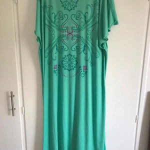 Turkos koftan med mönster - Säljer en turkos koftan med lila och rosa dekorativt mönster framtill. Klänningen har lös passform och korta ärmar, perfekt för en avslappnad stil. Materialet känns mjukt och luftigt.