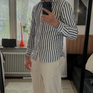 Randig skjorta från SMOG - Säljer en stilren skjorta från SMOG i vitt och mörkblått med vertikala ränder. Skjortan har lång ärm, klassisk krage och knäppning framtill. Perfekt för sommaren!☀️