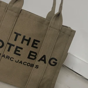  tote bag oliv grön - En olivgrön tote bag från Marc Jacobs med tydlig svart logga på framsidan. Väskan är i canvasmaterial och har två handtag. Perfekt storlek för att bära med sig det viktigaste och har en stilren desigin. Väldigt rymlig och perfekt söt storlek. Medföljer en bred rem för att bära över sig eller hängandes på axeln