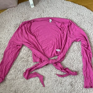 Rosa omlottopp med knyt - Säljer en rosa omlottopp med långa ärmar och knyt framtill. Perfekt att styla över ett linne eller klänning för en färgklick i garderoben. Tunn och mjuk i materialet, passar bra till vår och sommar.