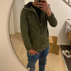 Olivgrön jacka med huva - Säljer en olivgrön jacka med huva och dragsko i midjan. Jackan har dragkedja och knappar framtill samt två stora fickor. Perfekt för kyligare dagar och har en stilren look.