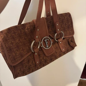 Brun mönstrad handväska från Ferre - Säljer en brun handväska från Ferre med snyggt geometriskt mönster och detaljer i syntetläder. Väskan har silverfärgade metalldetaljer, dubbla handtag och innerfack med dragkedja. Perfekt för dig som vill ha en unik accessoar.