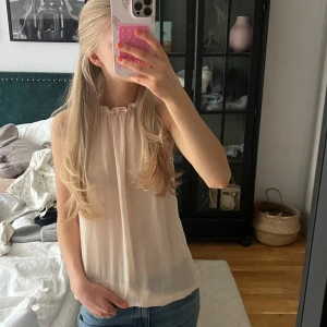 Ljusrosa ärmlös blus med rynkad hals - Säljer en söt ljusrosa/beige blus med rynkad halsringning och ärmlös design. Toppen har en lös passform och är tillverkad i ett tunt, lätt material. Det sitter en guldkedja i halsen så att toppen knäpps som ett halsband❣️