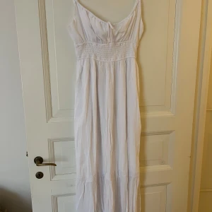 Vit långklänning från Hollister - Säljer en vit långklänning från Hollister med smala axelband och knytning i ryggen. Klänningen har smockad midja och är tillverkad i ett lätt, somrigt material. Perfekt för varma dagar och har en luftig passform.