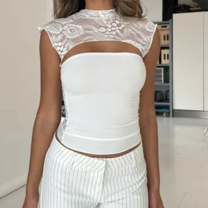Vit topp med spetsdetaljer - Säljer en vit topp med vacker spets över axlar och rygg. Toppen är ärmlös och har en tight passform med hög hals och fina blommiga spetsmönster. Perfekt för dig som vill ha en elegant och trendig look.