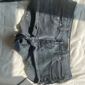 Svarta/gråa jeansshorts från One Teaspoon - Superkorta mörkblå jeansshorts från One Teaspoon med råa kanter och slitna detaljer. Klassisk femficksmodell med knapp och dragkedja framtill. Perfekta för varma dagar och en edgy stil.