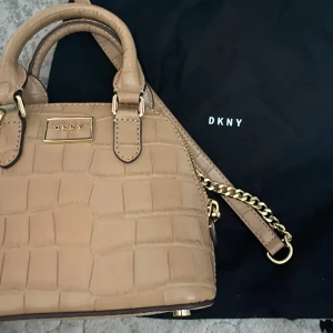 Beige handväska från DKNY med kedjedetalj - Säljer en beige handväska från DKNY med krokodilpräglat mönster och guldfärgad kedja. Väskan har både handtag och axelrem samt dragkedja upptill. Perfekt accessoar för att lyfta din outfit.