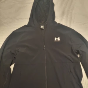 Svart jacka från Under Armour - Svart jacka från Under Armour med dragkedja och huva. Klassisk logga på bröstet och två sidofickor. 